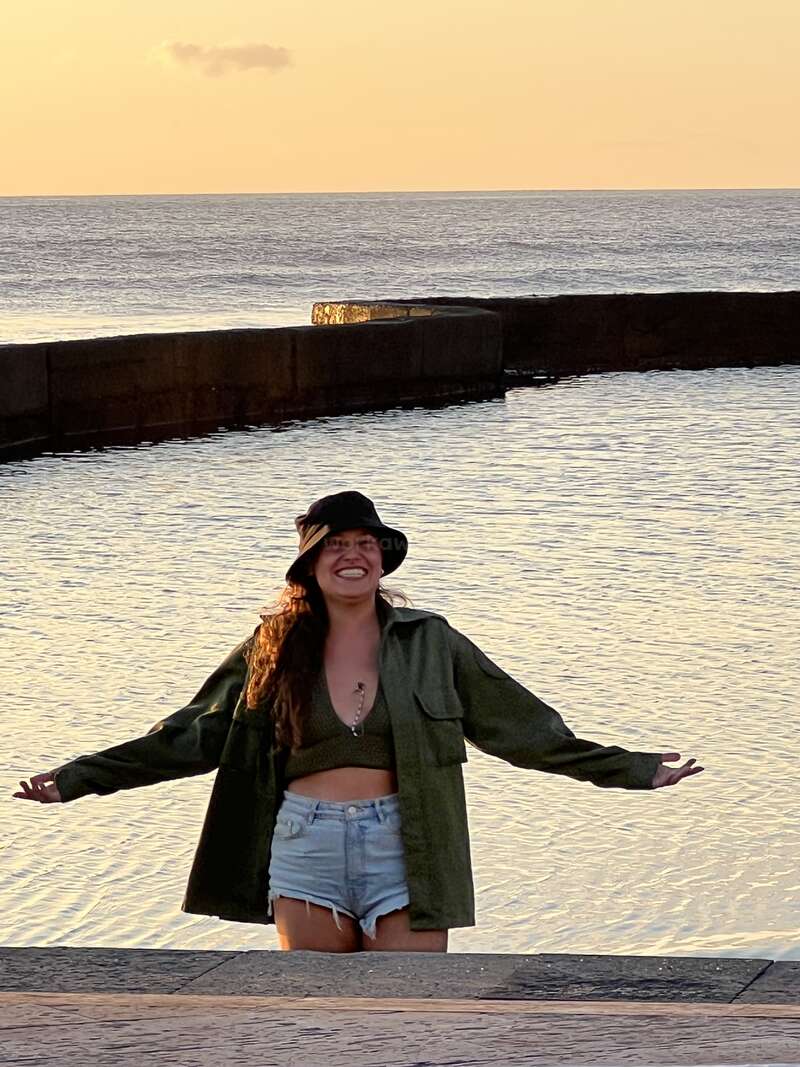 The image depicts a woman standing on a stone pier, arms outstretched, with her back to the ocean, wearing a hat, jacket, and shorts, in front of a calm sea at sunset.