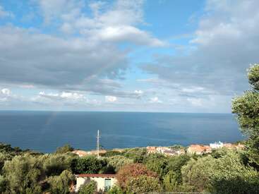 The image depicts a serene coastal town, with a cluster of houses nestled among lush trees, overlooking a vast expanse of calm blue water under a partly cloudy sky.