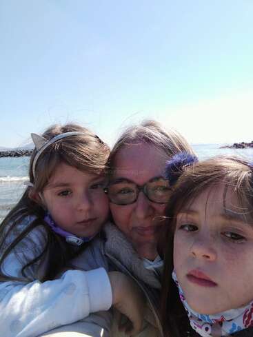 The image shows a woman and two young girls posing for a selfie on a beach, with the woman wearing glasses and the girls wearing headbands.