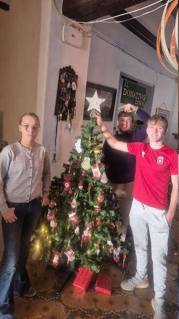 Trois personnes décorent un sapin de Noël festif avec des ornements et des lumières à l'intérieur. L'une d'elles place une étoile au sommet, tandis que les cadeaux sont disposés en dessous. La joie des fêtes emplit la scène.