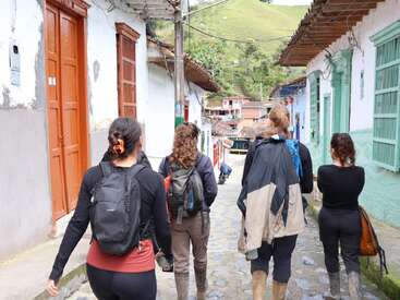 Cinq personnes marchent dans une rue pavée d'un village coloré, équipées de sacs à dos et de bottes, entourées de bâtiments blancs et verts, avec des collines verdoyantes visibles au loin.