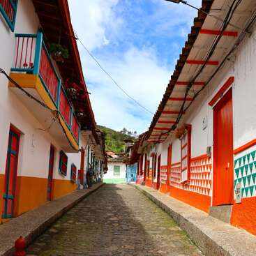 This image shows a charming cobblestone street lined with brightly colored houses, featuring red doors, balconies, plants, and vibrant geometric patterns under a blue sky.