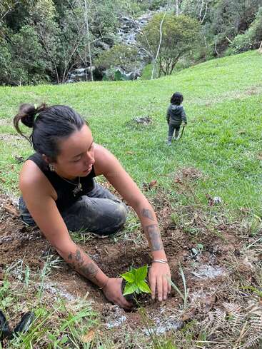 Une femme est agenouillée sur l'herbe et plante un petit jeune arbre. À l'arrière-plan, un enfant s'éloigne en direction des arbres et d'un ruisseau qui coule au milieu de la nature.