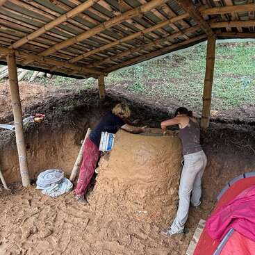 Deux personnes s'affairent à construire un four en terre ou une structure similaire sous un simple toit de bambou et de bois. Des palettes, une tente et des matériaux de construction sont éparpillés tout autour.