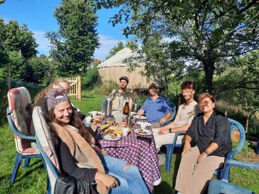 Um grupo de seis pessoas senta-se ao redor de uma mesa ao ar livre, saboreando alimentos e bebidas em um jardim verde e ensolarado, com árvores e uma iurta ao fundo.