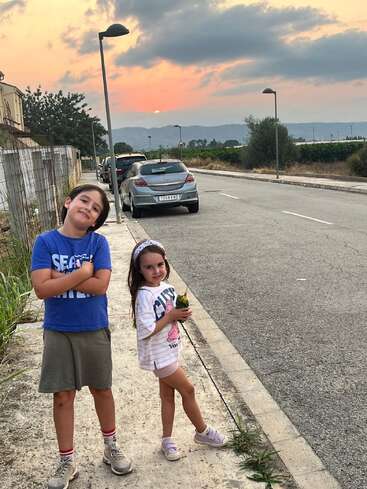 Duas crianças posam confiantes em uma rua tranquila de subúrbio ao pôr do sol, com carros estacionados, vegetação, montanhas e um céu dramático proporcionando um cenário lindo e sereno.