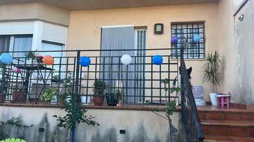 Esta imagem mostra uma varanda decorada com balões coloridos presos a uma grade de metal preta, vários vasos de plantas, degraus e uma pequena mesa com itens infantis.