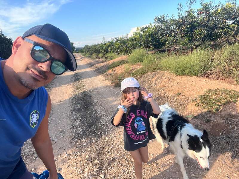 Um homem, uma menina e um cachorro preto e branco estão andando em um caminho de terra cercado por vegetação sob um céu azul claro e brilhante.