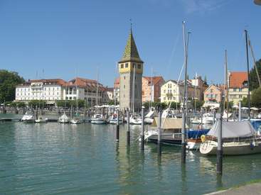 The image depicts a serene harbor scene with a prominent tower, featuring a row of boats docked in the water, surrounded by buildings and a clear blue sky.