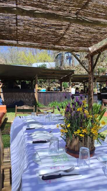 Uma mesa ao ar livre lindamente posta sob uma pérgula rústica, decorada com um arranjo floral vibrante no centro. A luz do sol penetra, criando sombras. Um ambiente descontraído de jardim com um bar nas proximidades.