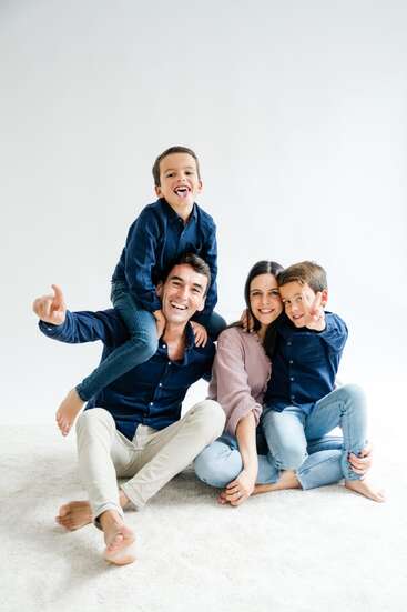 Una familia feliz de cuatro miembros, sonrientes y sentados en el suelo. El padre tiene a un hijo sobre los hombros, mientras la madre abraza a otro niño juguetón.
