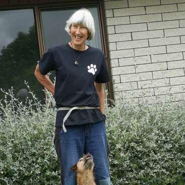A smiling woman with short gray hair stands outside by a brick wall and window, wearing a black shirt with a paw print, accompanied by a small dog.
