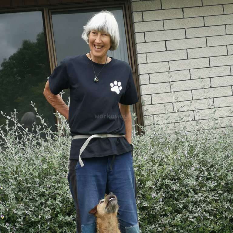 A smiling woman with short gray hair stands outside by a brick wall and window, wearing a black shirt with a paw print, accompanied by a small dog.