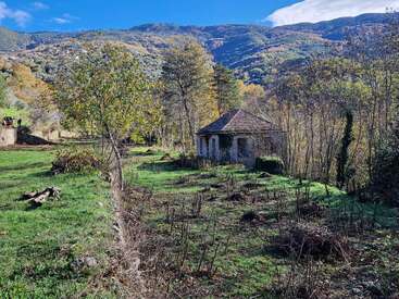 An old stone house stands alone among trees and green grass, surrounded by distant mountains under a blue sky with scattered clouds. Peaceful rural landscape.