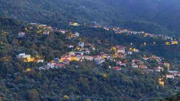 This image shows a picturesque hillside village at dusk, with scattered houses and buildings illuminated by warm lights, surrounded by lush green trees and dense forested mountains.