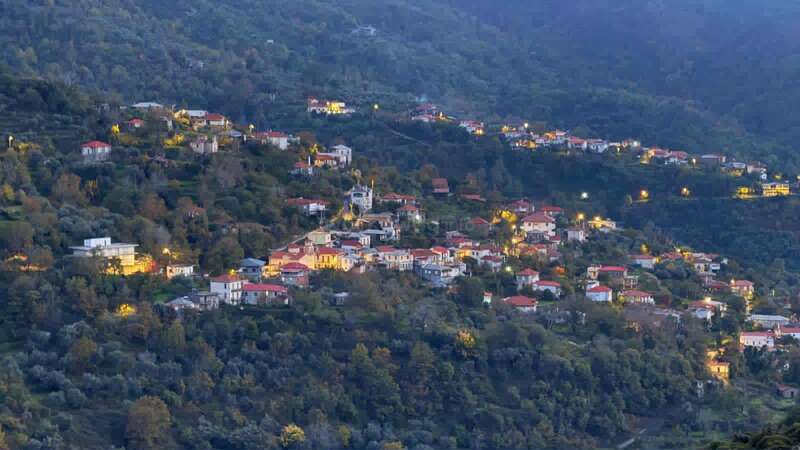 This image shows a picturesque hillside village at dusk, with scattered houses and buildings illuminated by warm lights, surrounded by lush green trees and dense forested mountains.