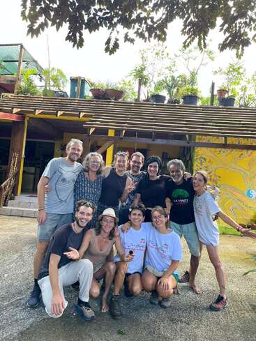 Un alegre grupo de doce personas posa frente a un edificio colorido con plantas en la azotea. Todos sonríen, se muestran relajados y disfrutan juntos de un día soleado y agradable.