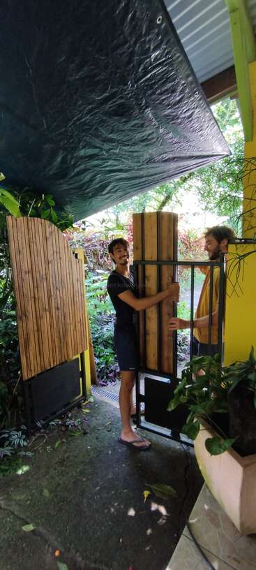 Dos hombres sonrientes trabajan juntos para instalar o reparar una verja de madera junto a una casa amarilla, rodeados de exuberantes plantas verdes y una lona negra que les da sombra.