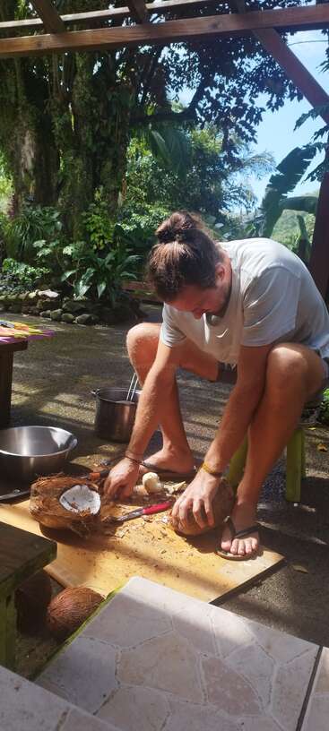 Un hombre está sentado al aire libre en un taburete pequeño, utilizando herramientas para abrir cocos sobre una tabla, rodeado de plantas tropicales, la luz del sol y cuencos de cocina.