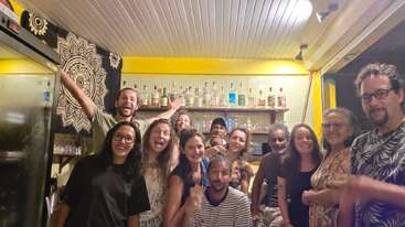 Un grupo de trece personas posa alegremente en un bar acogedor y con una iluminación cálida, con botellas en las estanterías a sus espaldas, sonriendo y disfrutando de una animada reunión social.