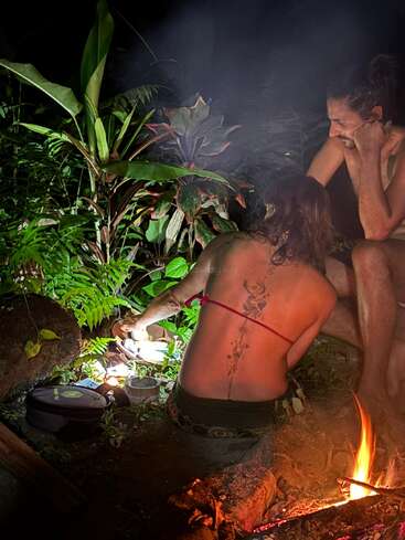 Dos personas están sentadas junto a una hoguera por la noche, rodeadas de una exuberante vegetación. Una de ellas tiene un gran tatuaje en la espalda. La escena transmite paz, calidez y cercanía a la naturaleza.