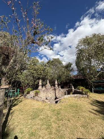 Uma cena de jardim exuberante com uma estrutura de pedra exclusiva, cercada por vegetação, árvores floridas, céu azul, nuvens espalhadas e assentos ao ar livre, criando uma atmosfera pacífica.