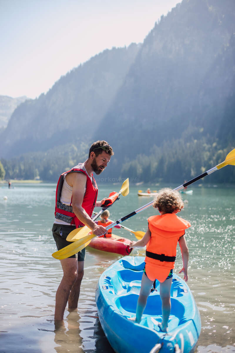 Un homme et un enfant se tiennent dans une eau peu profonde, tenant des pagaies et un kayak bleu, avec un autre kayak et des montagnes en arrière-plan, lors d'une journée ensoleillée.