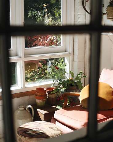 A cozy sunlit corner with a pink armchair, yellow cushion, potted plants, stacked terracotta pots, and a patterned ottoman beside a bright window.