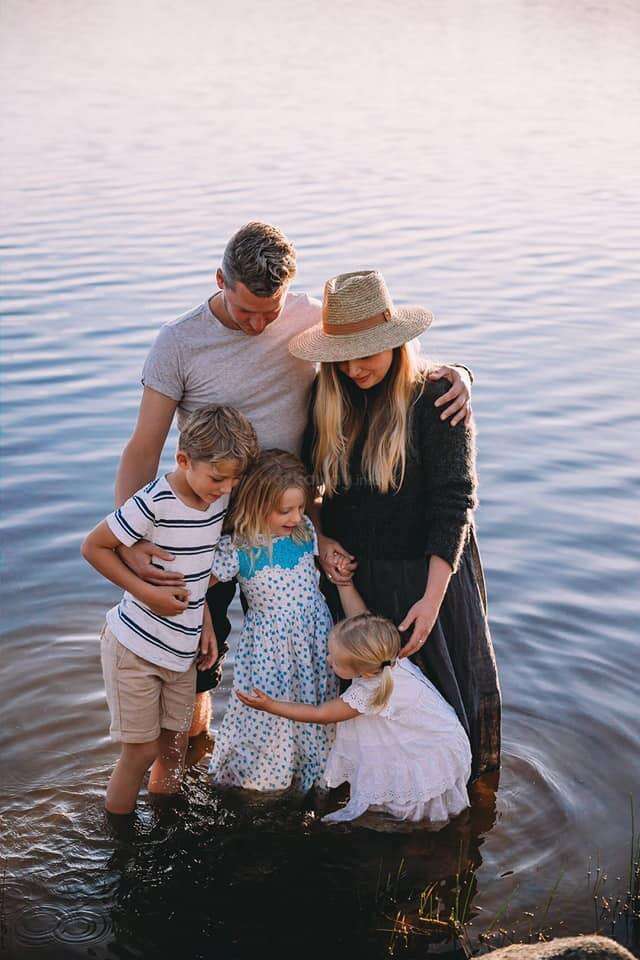 A family of five stands together in shallow water, sharing a tender moment. The mother wears a sunhat. Children smile, feeling the water around their feet.