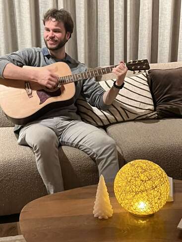 Un homme souriant est assis sur un canapé beige et joue de la guitare acoustique. Un éclairage chaleureux, une lampe sphérique brillante et une bougie blanche décorative créent une atmosphère confortable.