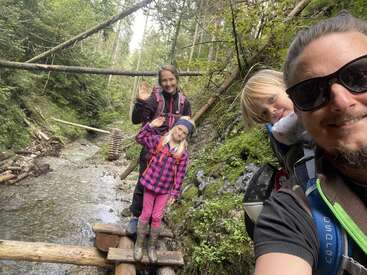 Une famille souriante de quatre personnes se promène dans une forêt luxuriante et traverse un pont en bois au-dessus d'un ruisseau. Ils sont habillés pour l'aventure, font des signes de la main et profitent ensemble de la nature.