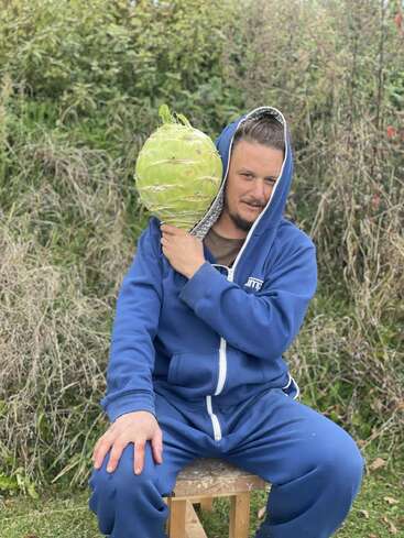 Un homme vêtu d'un survêtement bleu est assis sur un tabouret en plein air et s'amuse à tenir un gros chou vert près de sa tête, imitant ainsi un téléphone.