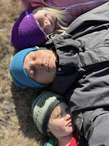 Un homme et deux enfants, tous coiffés de chapeaux colorés et vêtus de vestes chaudes, sont allongés ensemble sur le sol, les yeux fermés, profitant paisiblement d'un moment ensoleillé en plein air.