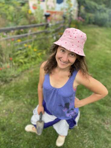 Une femme souriante portant un chapeau rose et un débardeur violet se tient à l'extérieur sur de l'herbe, tenant une truelle de jardinage. Des fleurs éclatantes et une clôture décorative se trouvent derrière elle.