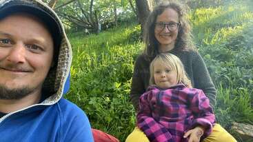 Un homme, une femme et un jeune enfant sont assis sur l'herbe, souriants. Les arbres et la lumière du soleil les entourent. La scène donne une impression de chaleur, de joie et de détente dans la nature.