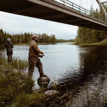 Das Bild zeigt zwei Männer, die unter einer Brücke angeln. Der eine steht auf einem Felsen im Wasser und hält eine Angelrute, der andere steht am Flussufer.