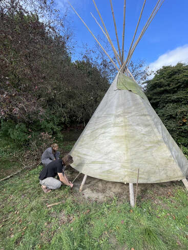 Two people are setting up a tipi in a grassy area, surrounded by trees. The tipi is covered in a translucent material and wooden poles extend upward.