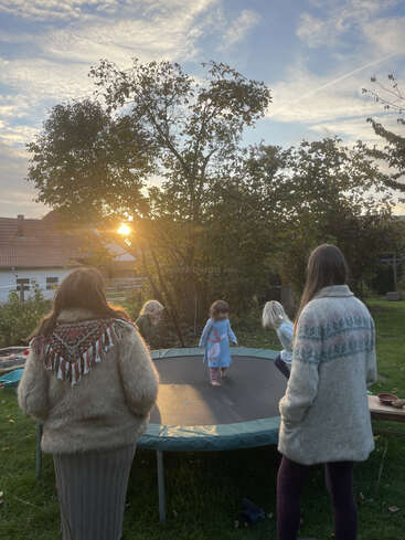Two adults stand beside a trampoline, watching three young children play. The scene is set in a backyard during sunset, with trees and houses in view.