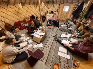 A group of people sit in a cozy yurt, gathered in a circle. They are working with papers, sharing food, and engaging in group discussion.