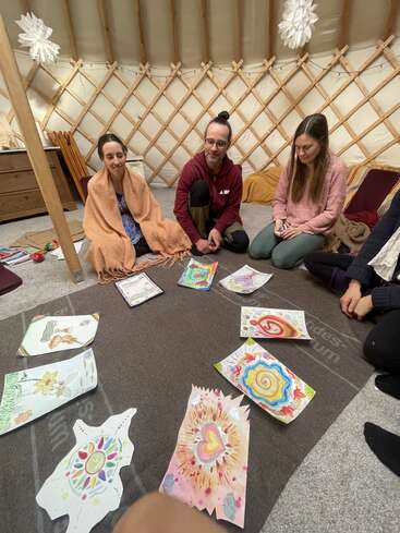 Four people sit on the floor of a cozy yurt, surrounded by colorful, abstract artwork laid out on a rug, sharing creative expressions in a circle.