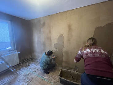 Two people are plastering and repairing a wall in a room. The floor is covered for protection, and there are work tools and a window visible.