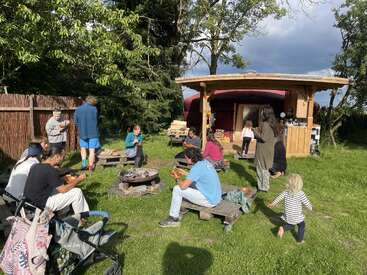 La imagen muestra a un grupo de personas reunidas en una zona de césped, con una estructura de madera y una hoguera al fondo, rodeadas de árboles y un cielo nublado.