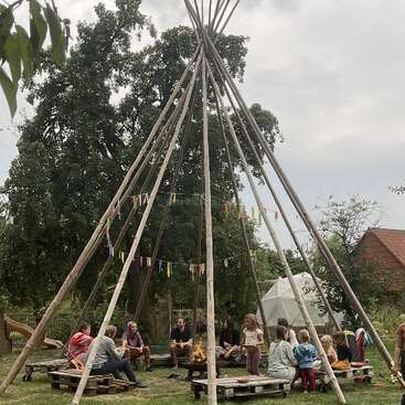 Eine Gruppe Leute, darunter auch Kinder, sitzt draußen unter einem großen, tipiartigen Holzbau um ein Lagerfeuer herum, umgeben von Bäumen und Grün, und quatscht gemütlich.