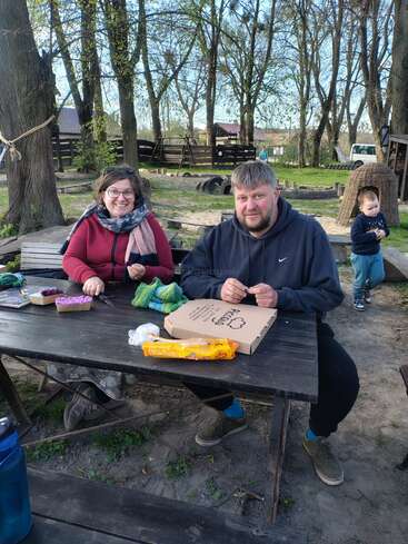 Ein Mann und eine Frau sitzen draußen an einem Picknicktisch, lächeln und basteln. Auf dem Tisch stehen Lebensmittel und eine Pizzaschachtel. Im Hintergrund spielt ein Kind.