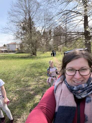 Eine Frau macht draußen ein Selfie – sie trägt eine Brille und einen Schal. Hinter ihr laufen Kinder auf einem grasbewachsenen, von Bäumen gesäumten Weg unter blauem Himmel.