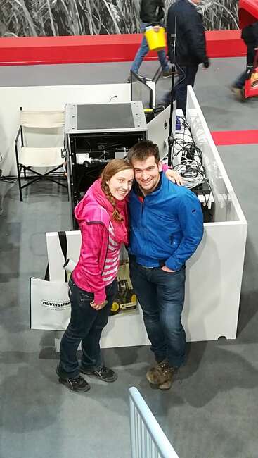 A smiling couple poses together indoors, standing beside a booth with electronic equipment. Both wear casual jackets. People and booths can be seen in the background.