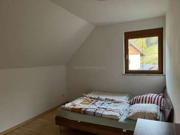 This is a simple, minimalistic bedroom with a double bed, patchwork blankets, and striped pillows. A single window lets in natural light, revealing countryside views.