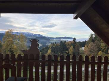 Un acogedor balcón con vistas a un pintoresco valle, rodeado de árboles otoñales y lejanas montañas nevadas. Una valla de madera con una bonita decoración con siluetas de vacas.