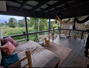 A cozy covered porch features a bed with pillows, hammocks, rocking horse, trampoline, chairs, tables, a lush green view, and relaxing, rustic decor.