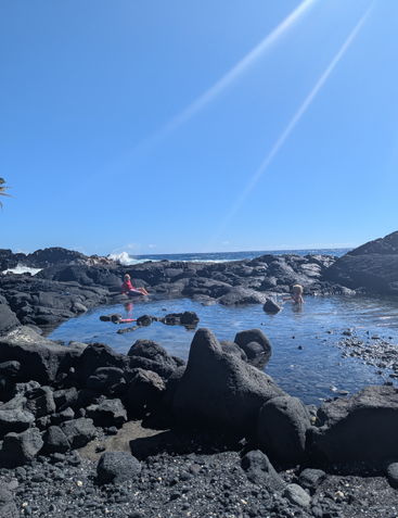 Two children play in a natural tidal pool surrounded by black volcanic rocks under a bright blue sky, with sunshine and ocean waves in the background.